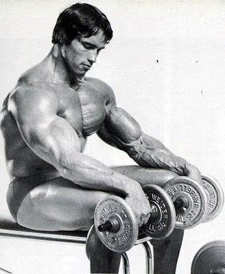 Black-and-white photo of a heavily muscled bodybuilder sitting and reaching down to grip a row of dumbbells lined up on a bench.