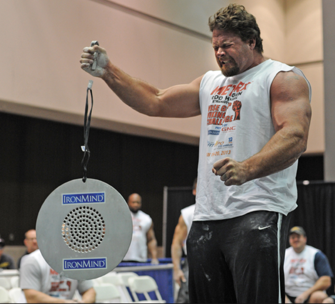 A bearded, muscular strongman competitor in a white event tank top grips a small device attached by a strap to a large round perforated metal plate branded "IronMind," gritting his teeth with a clenched fist, at an indoor strength competition with spectators seated behind him.