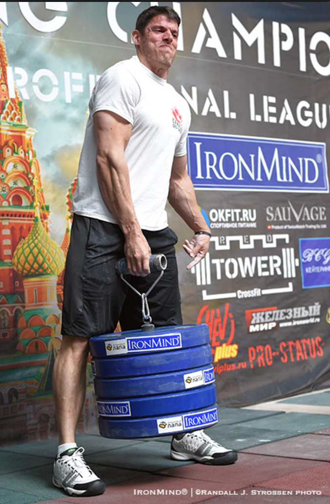 A strongman competitor in a white t-shirt and shorts holds a thick rotating handle attached to a stack of blue IronMind weight plates, in front of an event backdrop featuring Saint Basil's Cathedral and sponsor logos including IronMind, at a Moscow strongman grip competition.