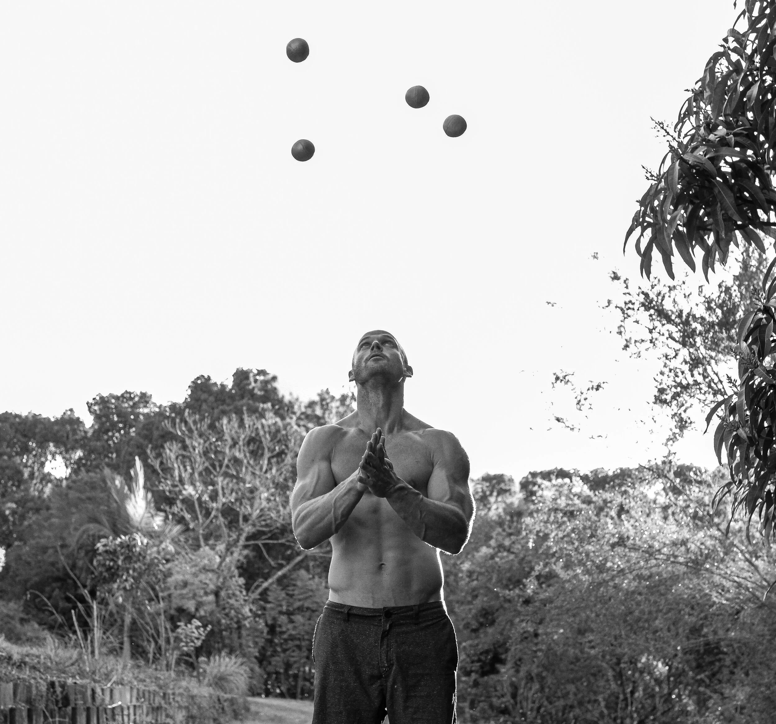 Black-and-white photo of a shirtless, muscular bald man juggling five balls outdoors, photographed from a low angle against the sky with trees in the background.