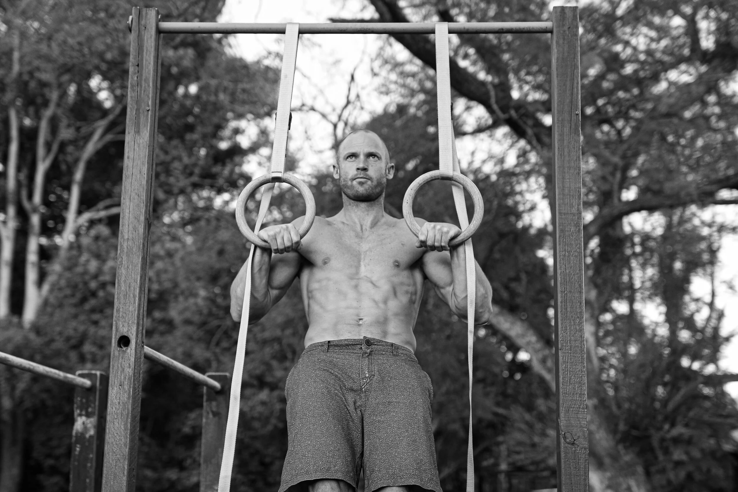 Black-and-white photo of a bearded, muscular bald man holding gymnastic rings during a dip, suspended from an outdoor pull-up rig with trees in the background.
