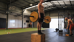 A shirtless man standing on a wooden box performs a Jefferson curl with a barbell in a warehouse-style gym, with other people visible in the background.