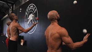 Two shirtless men juggling balls to each other indoors in front of a gym wall with a circular logo.