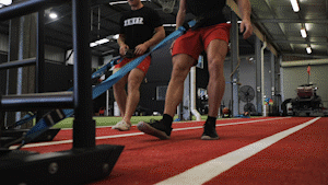Two people in red shorts pulling a weighted sled across a red turf gym floor.