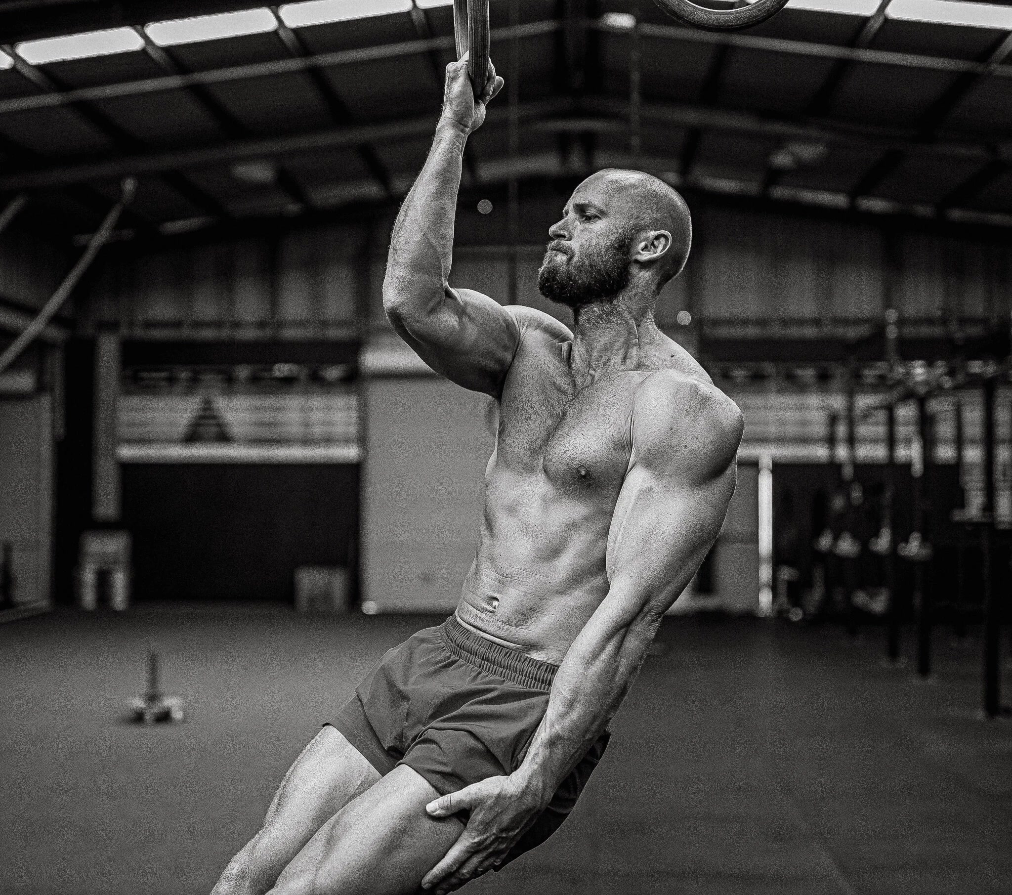Black-and-white photo of a bearded, muscular bald man holding onto gymnastic rings mid-pull in a warehouse-style gym.