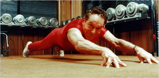 An older muscular man in a red long-sleeve top and shorts performs a push-up balanced on his fingertips on a carpeted floor, with racked dumbbells lining the wall behind him in a home gym.