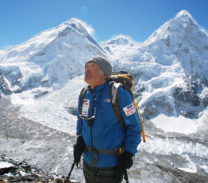 A mountaineer in a blue jacket, backpack, and beanie stands looking upward, with snow-capped mountain peaks in the background.