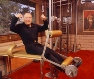 An elderly man in a black tracksuit sits on gym equipment, smiling and flexing both biceps, in a home gym with red carpet and wood-paneled walls.